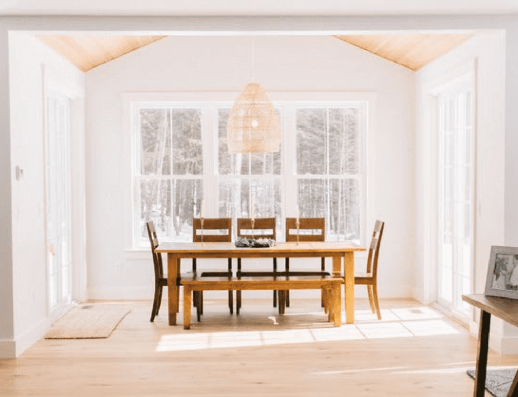 A modern dining room with a wood ceiling and floors, white walls, dining room table that seats eight and three double hung windows on the back wall.