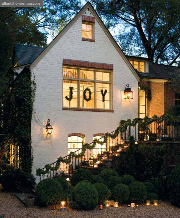 This white, brick house has garland over the banister, candles by the bushes and “JOY” spelled out using wreaths over the windows. 