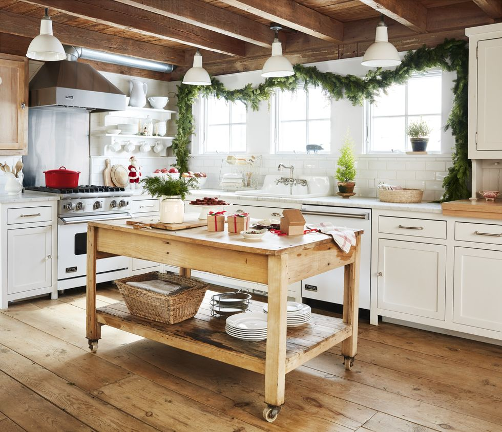 This large country kitchen has green garland hanging across three windows above the sink. The rolling island is holding some kitchen items, including plants, baskets, a cutting board and a dishtowel.
