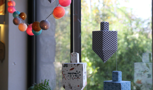Small dreidels are hanging in a window. One has a blue chevron pattern, one has a light blue design and the other has white, green and red patterns. 