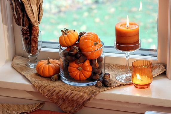 Alt Text: This window sill is featuring two orange candles, a jar of pumpkins and acorns, a glass of corn and a burlap cloth.