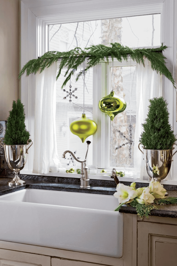 This kitchen design has a white farmhouse sink and a dark marble countertop. There are big ornaments hanging in front of the kitchen window, two small trees in silver vases on the sides of the sink and some garland draped across the window. 