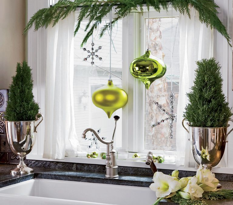 This kitchen design has a white farmhouse sink and a dark marble countertop. There are big ornaments hanging in front of the kitchen window, two small trees in silver vases on the sides of the sink and some garland draped across the window.