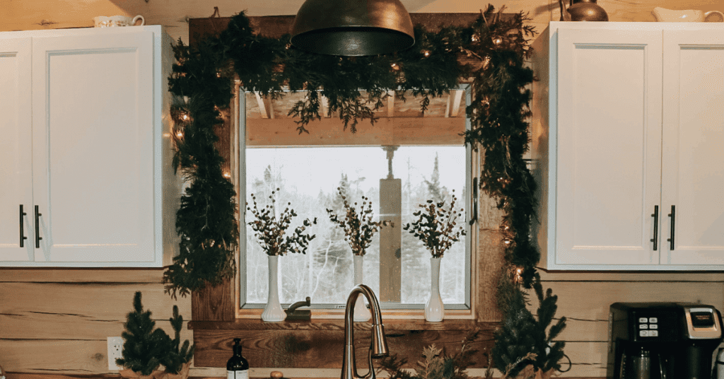 This kitchen has a window above the sink. There is garland hanging above and around the window, and there are three white vases on the window sill holding seasonal foliage. There are white cabinets on each side of the sink. 