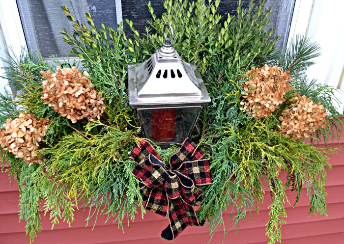This red house has a flower box below the window that’s decorated with Christmas decor. There is greenery, dried hydrangeas, a metal lantern and a plaid bow. 