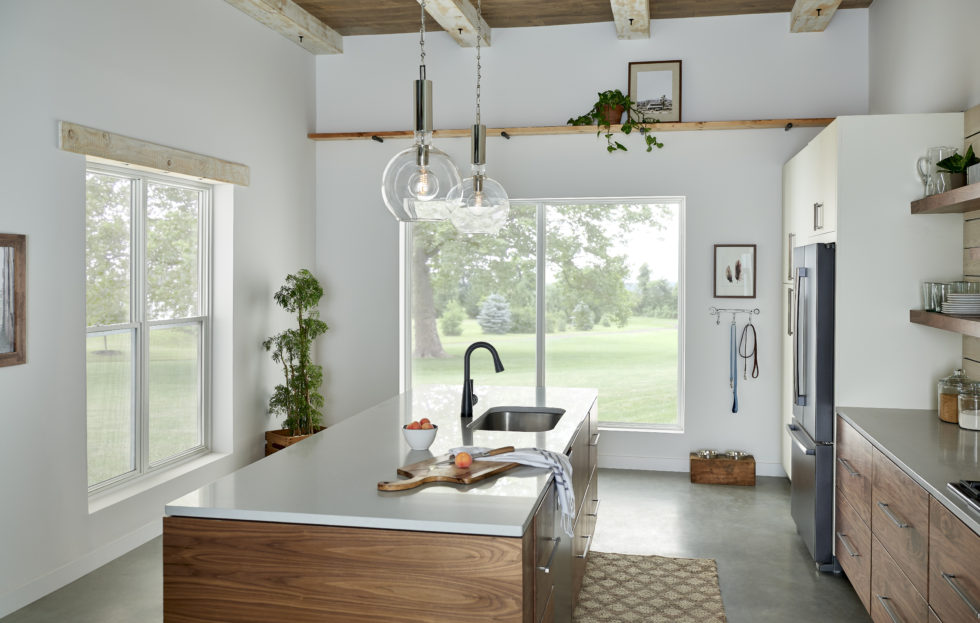 Alt text: A kitchen with an island, hanging light fixtures, wooden beam ceiling, a house plant, a patterned rug, dog leashes hanging on the wall and oranges on the counter. There are two picture windows on the far wall, and two double-hung windows on the left wall.